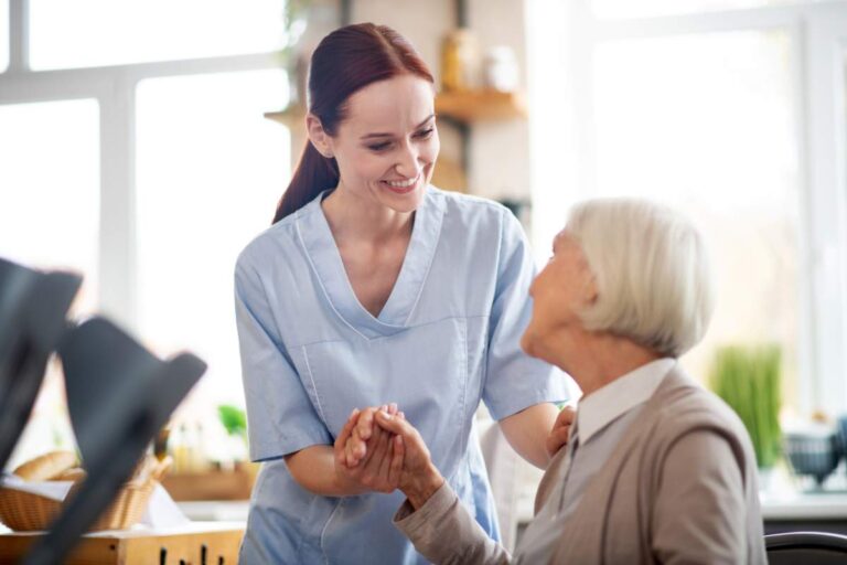 An older adult and a family caregiver sitting together at a kitchen table, sharing conversation and a warm drink.