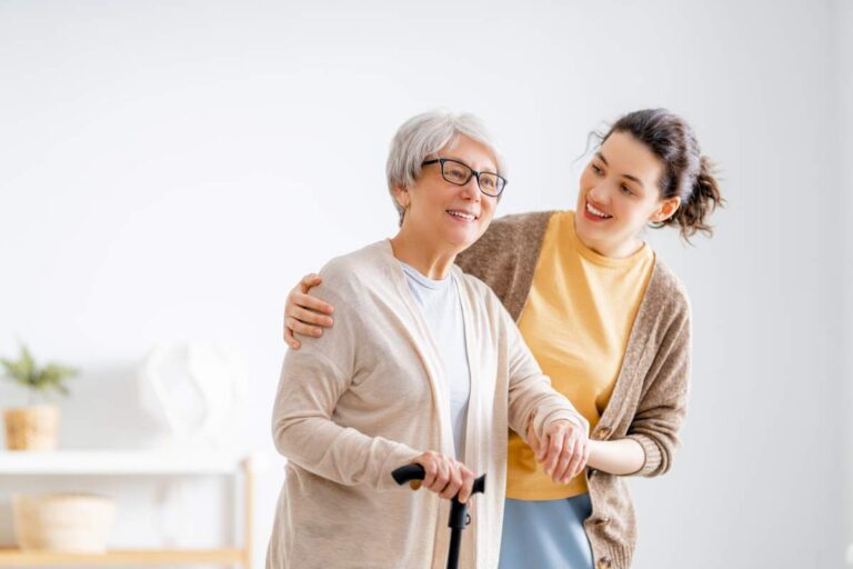 An elderly person and a middle-aged adult sit on a porch, talking gently as they hold hands.